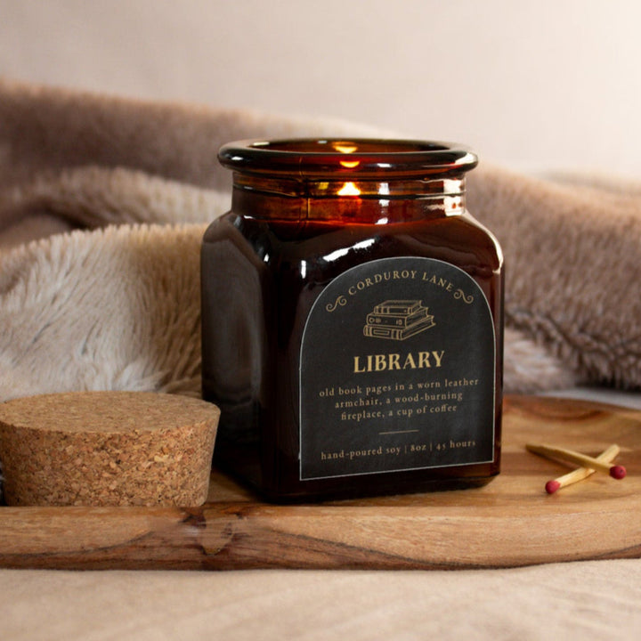 Candle in an amber jar labeled 'Library' on a wooden tray with a soft blanket in the background.