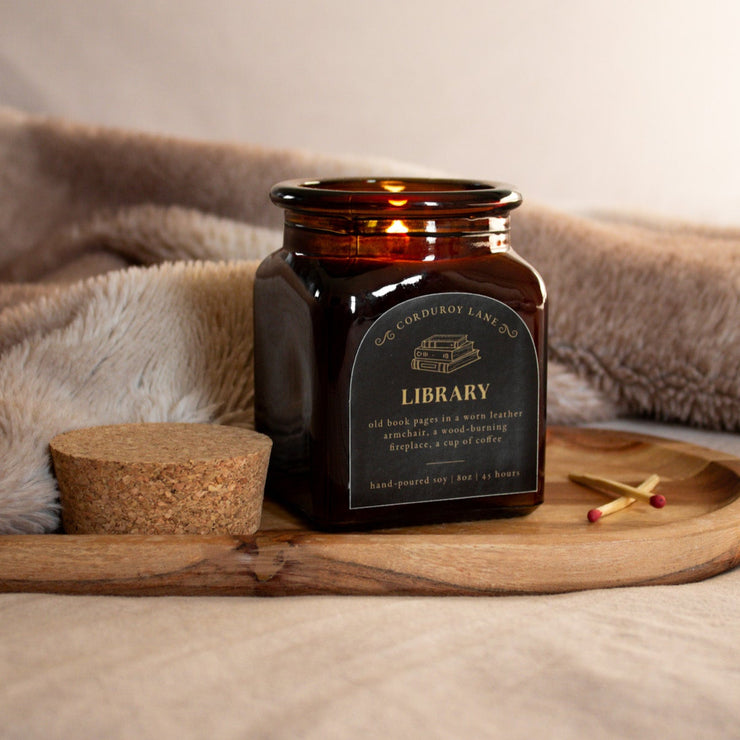 Candle in an amber jar labeled 'Library' on a wooden tray with a soft blanket in the background.