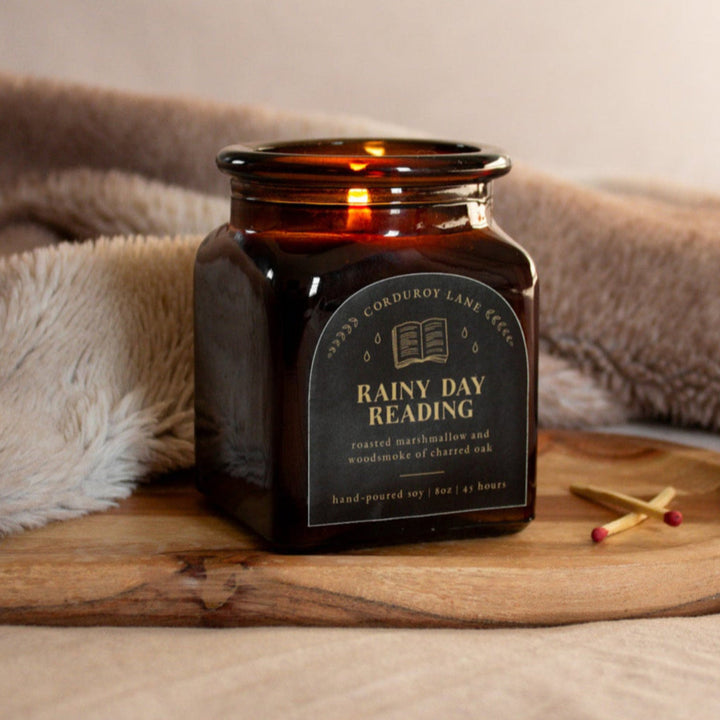Candle labeled 'Rainy Day Reading' on a wooden tray with a blanket in the background
