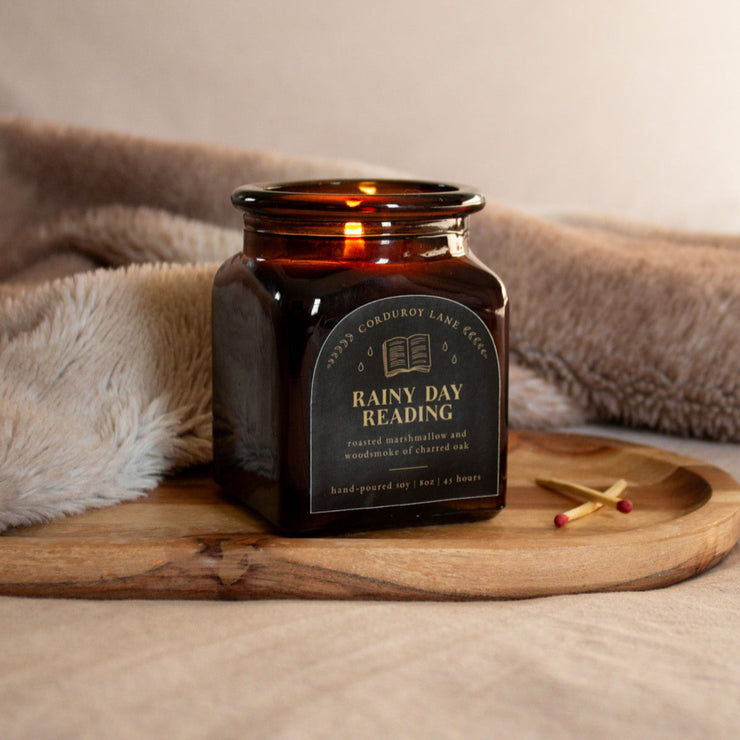 Candle labeled 'Rainy Day Reading' on a wooden tray with a blanket in the background