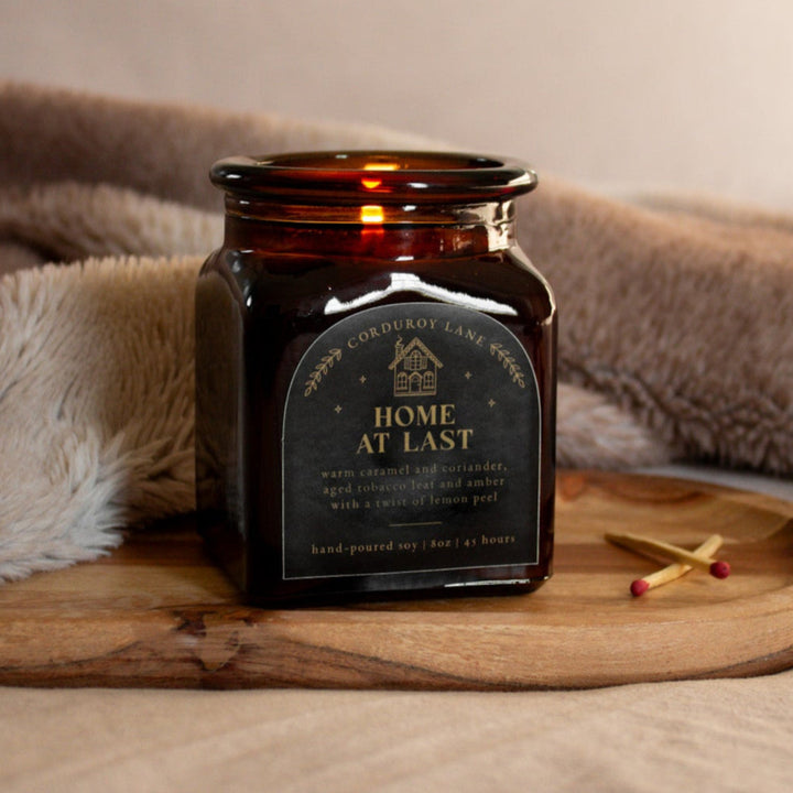 Home at Last: a comforting and cozy scented candle from Corduroy Lane. Warm caramel and coriander, aged tobacco leaf and amber with a twist of lemon peel. An amber glass square jar with a black and gold label and a cork lid, comes in a black gift bag