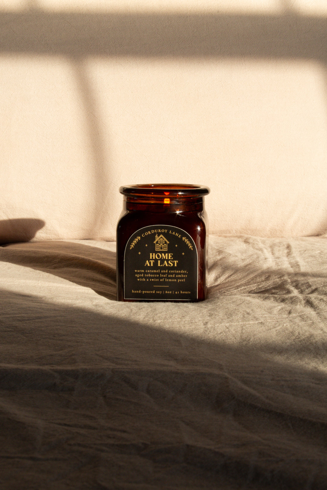 Home at Last: a comforting and cozy scented candle from Corduroy Lane. Warm caramel and coriander, aged tobacco leaf and amber with a twist of lemon peel. An amber glass square jar with a black and gold label and a cork lid, comes in a black gift bag