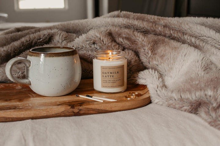 Candle labeled 'Oatmilk Latte' and a mug on a wooden tray with a blanket in the background