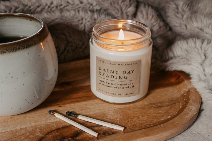 Candle labeled 'Rainy Day Reading' on a wooden surface with a mug and matches.