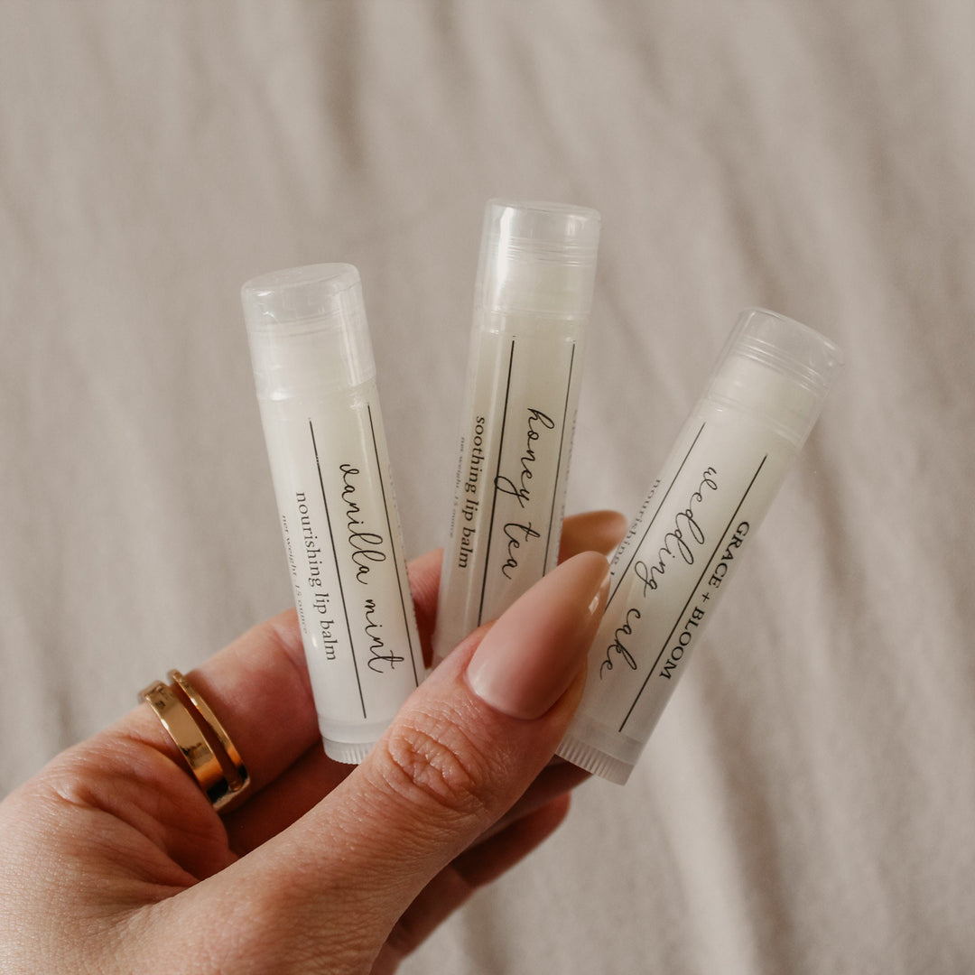 A hand holding three lip balm sticks with clear caps, labeled with different flavors such as 'Wedding Cake', 'Birthday Cake', and 'Honey Tea'.