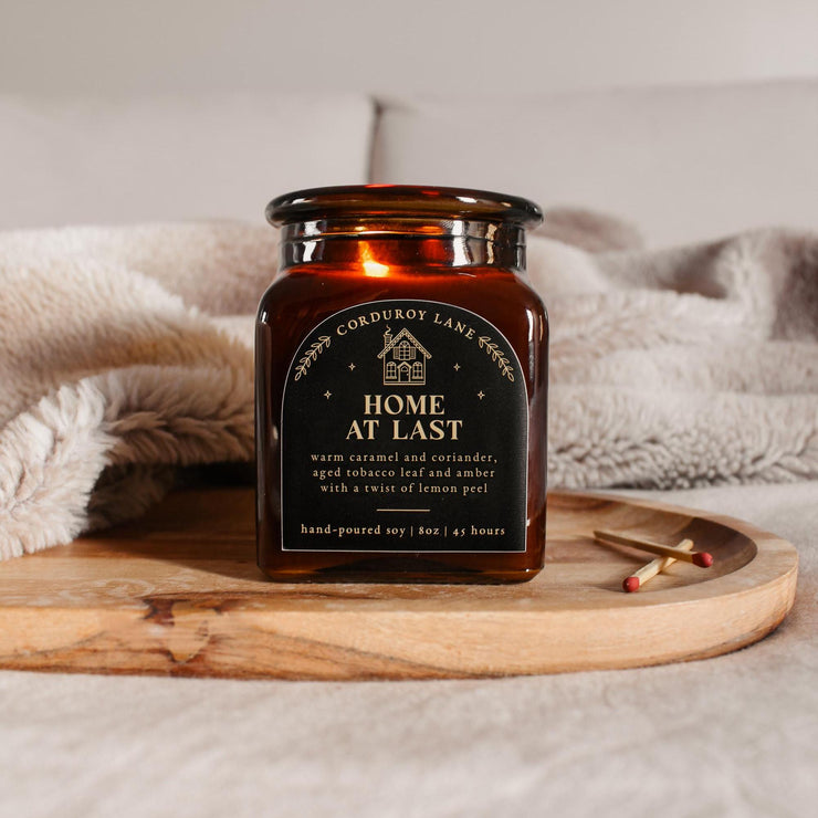 Home at Last: a comforting and cozy scented candle from Corduroy Lane. Warm caramel and coriander, aged tobacco leaf and amber with a twist of lemon peel. An amber glass square jar with a black and gold label and a cork lid, comes in a black gift bag