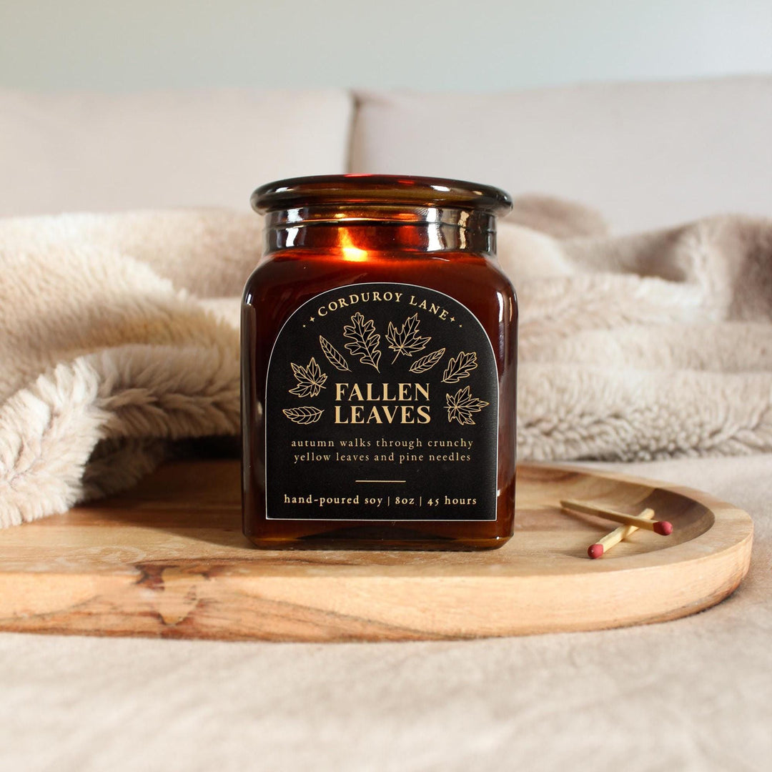 Square amber candle jar labeled 'Fallen Leaves' on a wooden tray with a soft blanket in the background