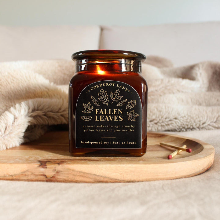 Square amber candle jar labeled 'Fallen Leaves' on a wooden tray with a soft blanket in the background
