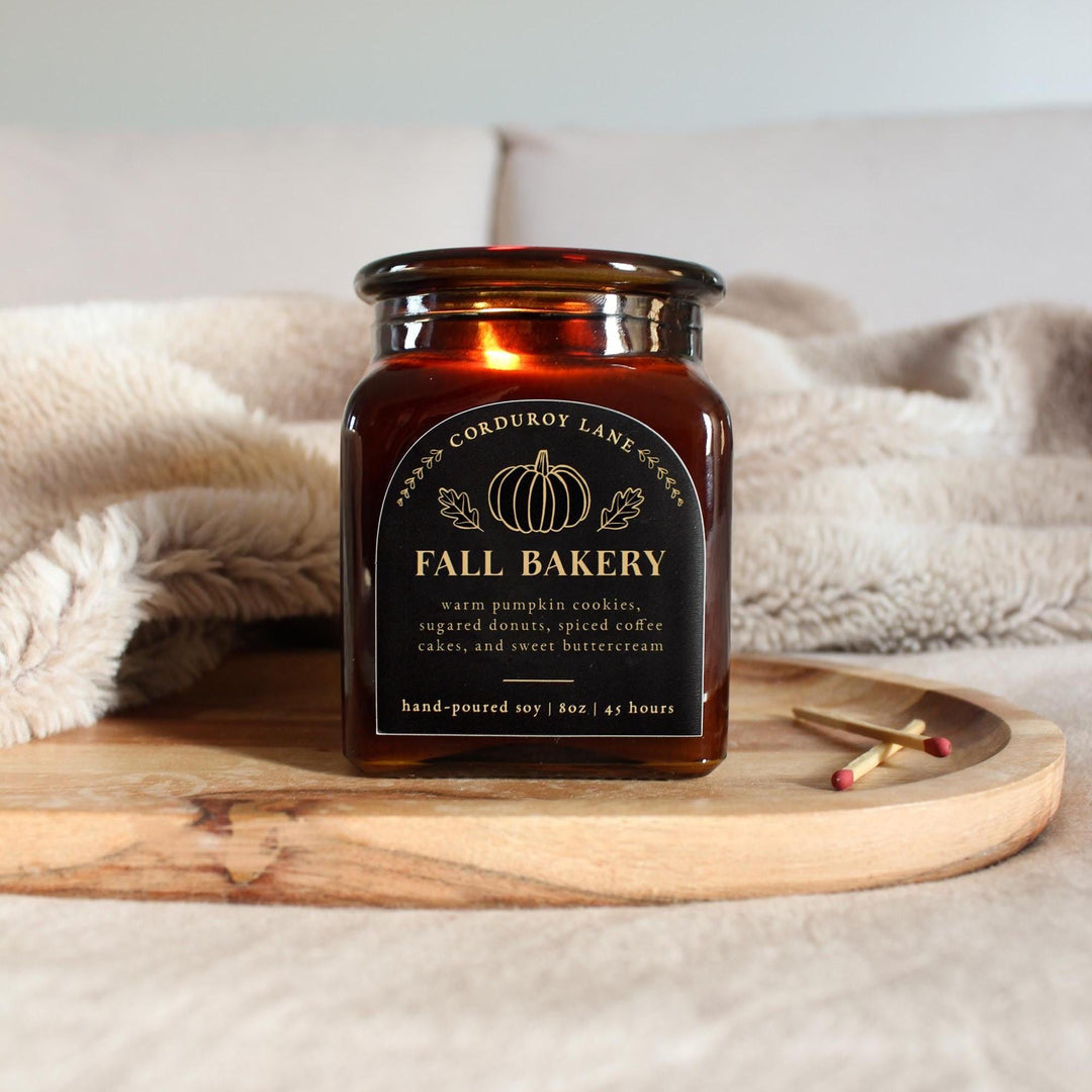 Candle jar labeled 'Fall Bakery' on a wooden tray with a soft blanket in the background