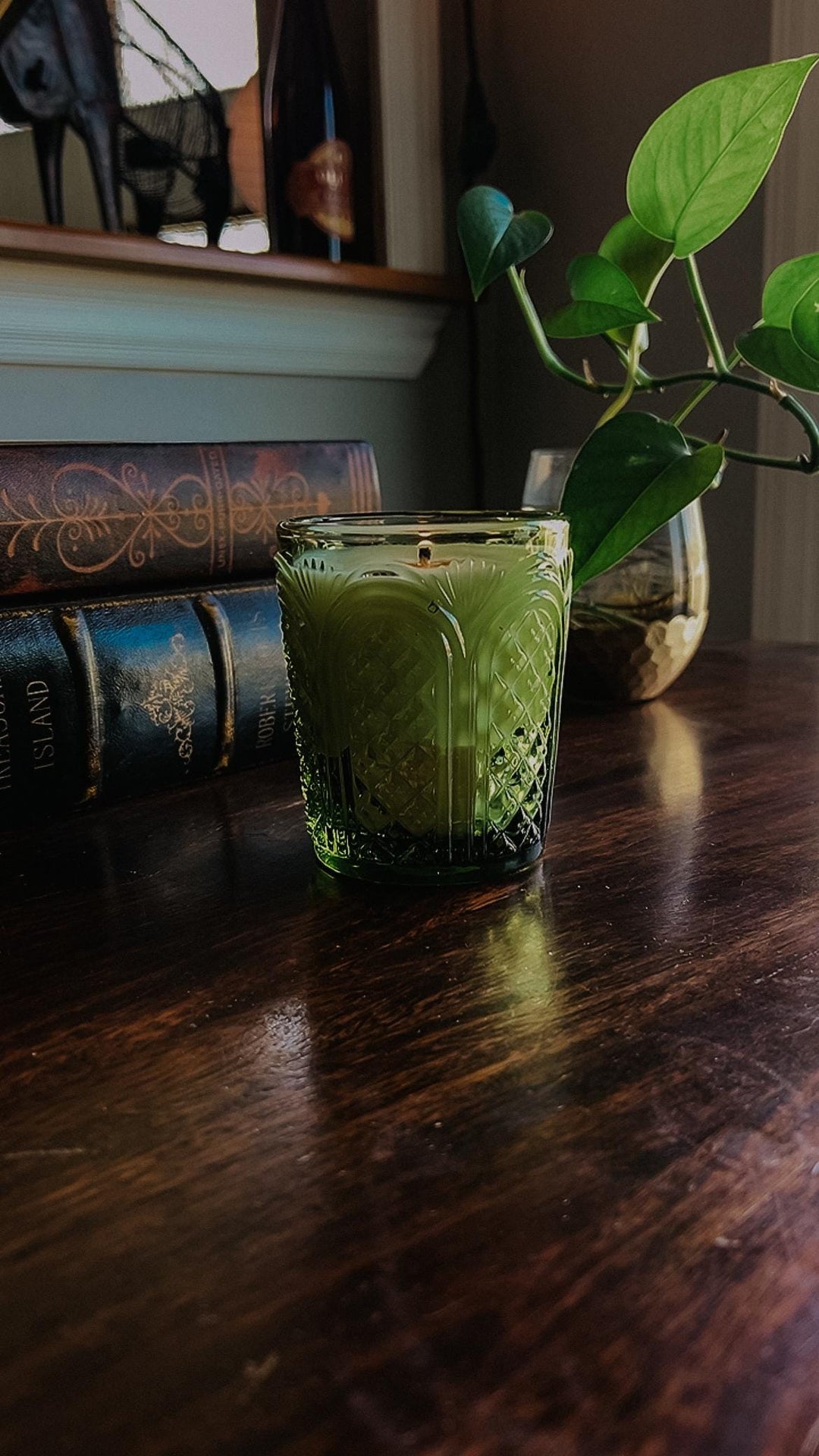 Green candle in a glass holder on a wooden surface with books and a plant in the background.