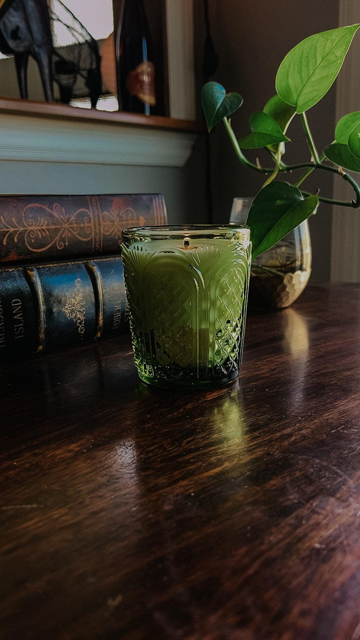 Green candle in a glass holder on a wooden surface with books and a plant in the background.