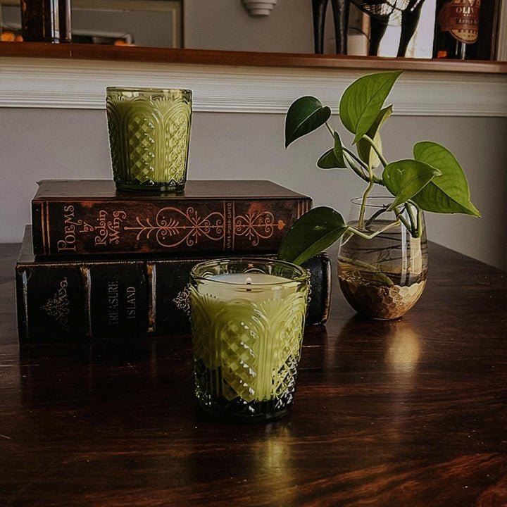 Green textured candles in a glass holder on a wooden surface with books and a plant in the background.