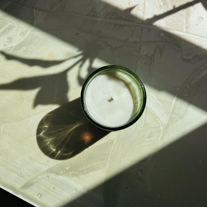 Candle in a glass container on a textured surface with leaf shadows
