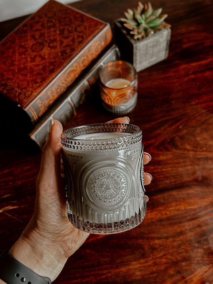 Hand holding a textured glass Speakeasy candle on a wooden surface with books and a plant in the background.