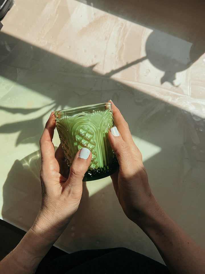 Person holding a Garden Mint candle in a green glass jar with intricate patterns on a light-colored surface.