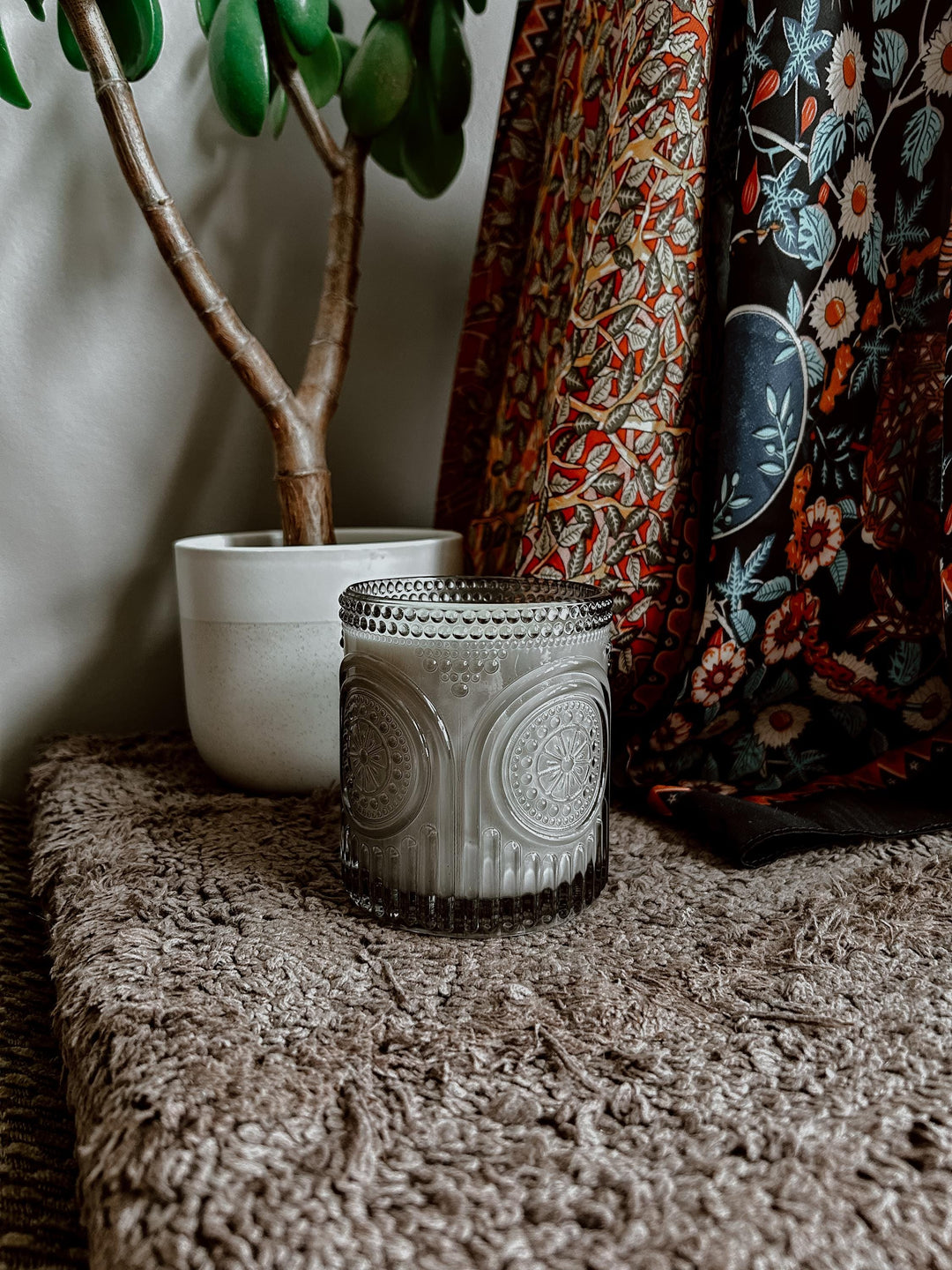 Decorative candle on a textured surface with a plant and patterned fabric in the background