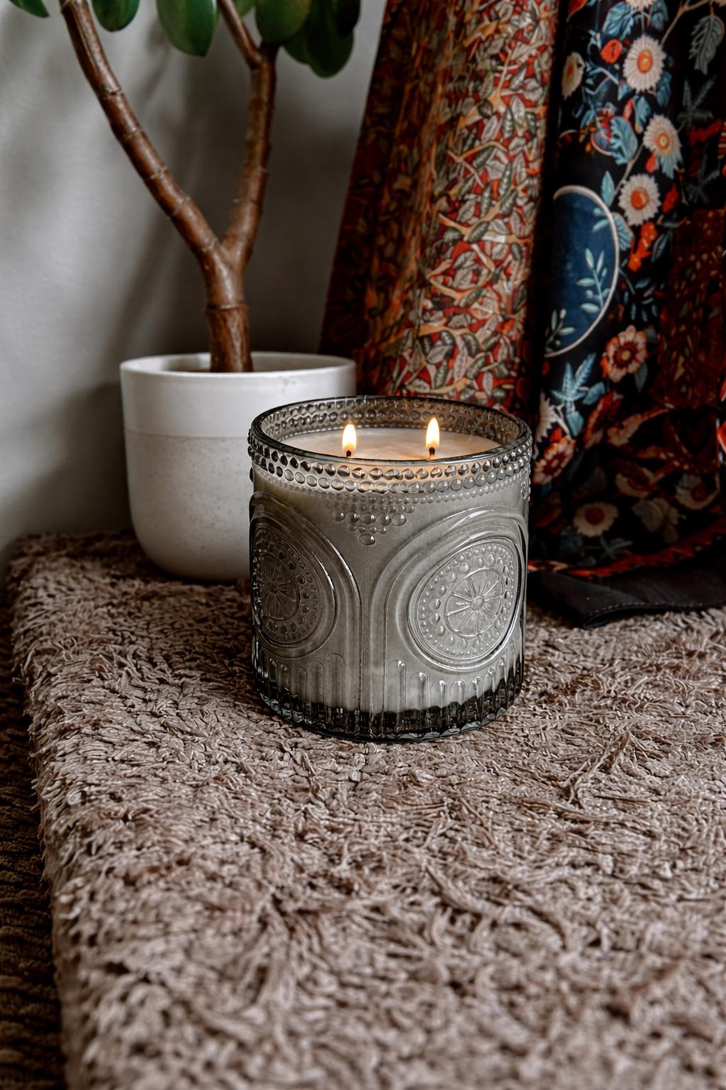 Decorative candle in a textured holder on a textured surface with a plant and patterned fabric in the background.