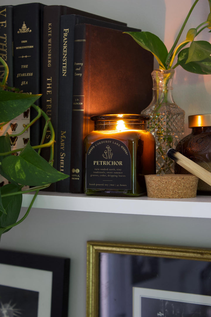 A lit, square green glass candle jar with a black label that reads "PETRICHOR" and "CORDUROY LANE". The candle is next to a stack of books and a glass vase with a plant. The candle is hand-poured soy, 8 oz, and burns for 45 hours.