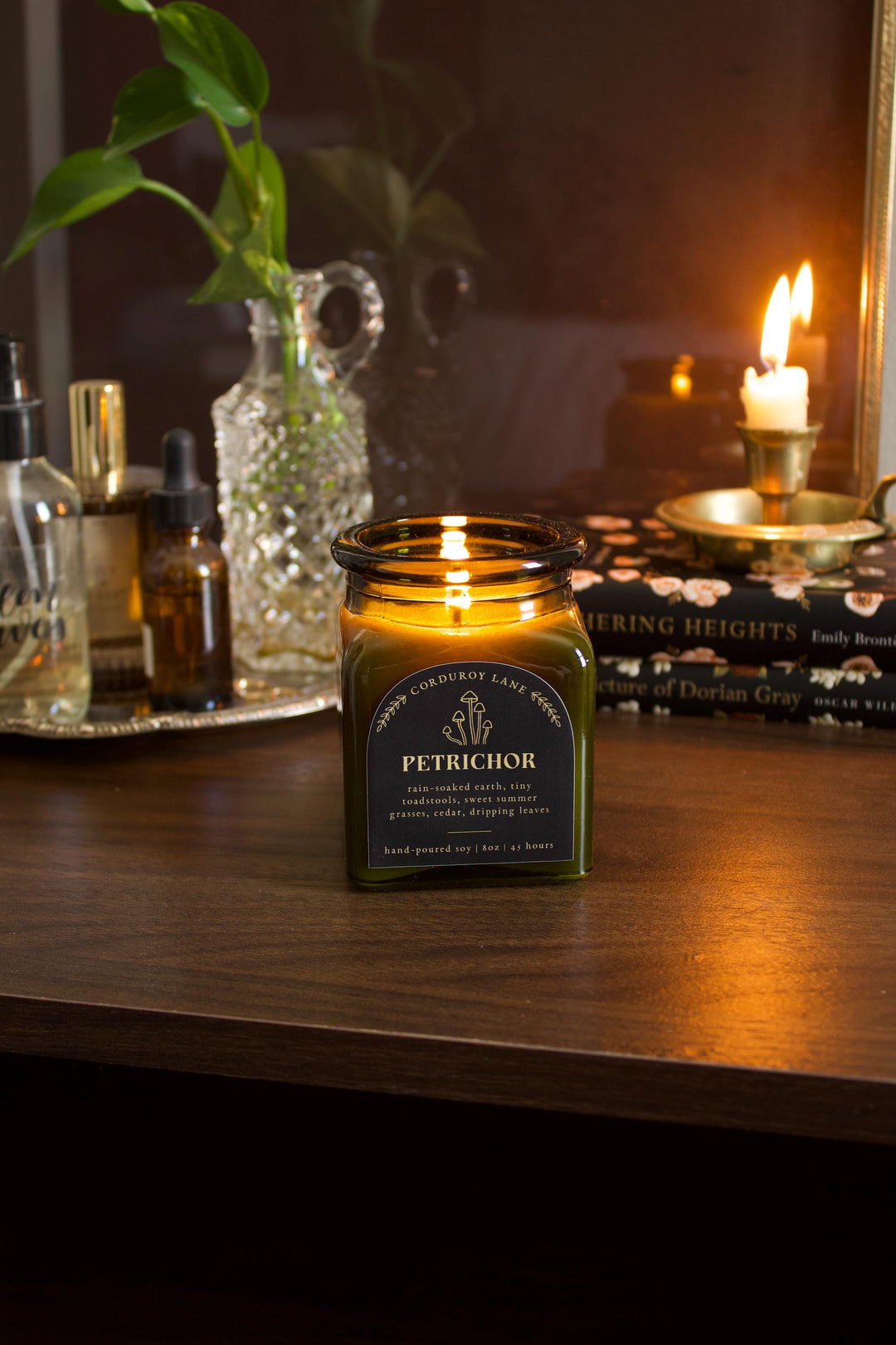 Candle in a jar labeled 'Petrichor' on a wooden surface with books and a plant in the background.