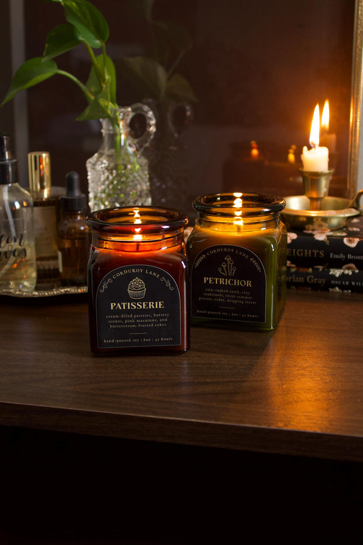 Two lit candles on a wooden surface with decorative bottles and a plant in the background.