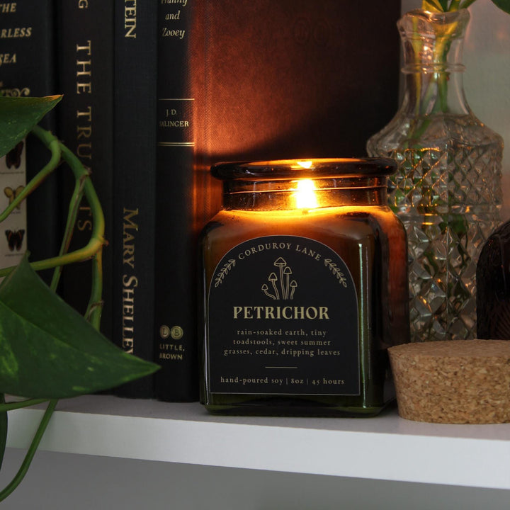 A lit, square green glass candle jar with a black label that reads "PETRICHOR" and "CORDUROY LANE". The candle is next to a stack of books and a glass vase with a plant. The candle is hand-poured soy, 8 oz, and burns for 45 hours.