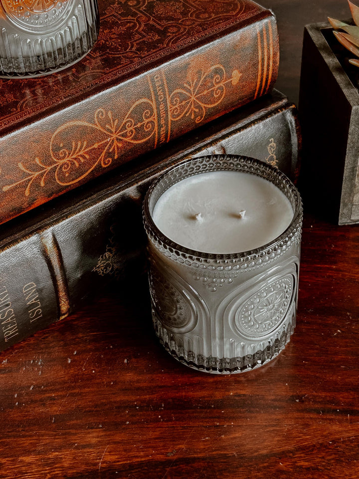 A gray, patterned glass candle holder with a white candle inside. The candle has two wicks. The candle holder is sitting on a dark wooden surface next to a stack of antique books.
