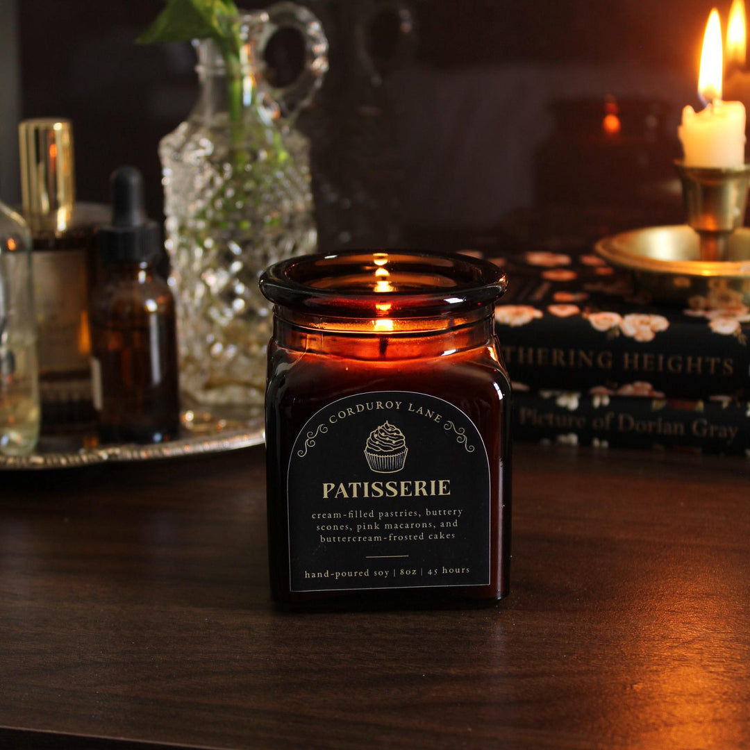 A lit, amber glass candle jar with a black label that reads "PATISSERIE" with a cupcake graphic. The candle is hand-poured soy and has a burn time of 45 hours. The candle sits on a dark wooden surface.