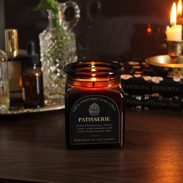 A lit, amber glass candle jar with a black label that reads "PATISSERIE" with a cupcake graphic. The candle is hand-poured soy and has a burn time of 45 hours. The candle sits on a dark wooden surface.