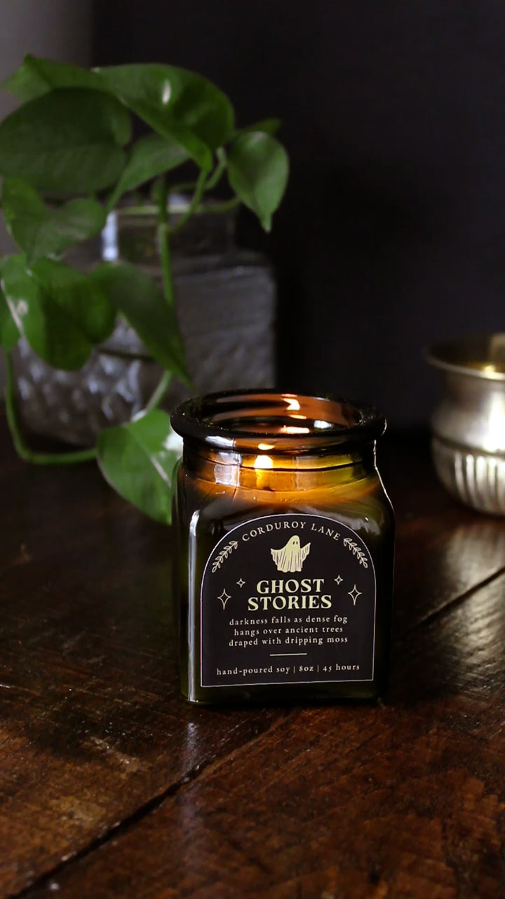 Ghost Stories: a spooky halloween candle from Corduroy Lane in a green glass square apothecary jar with a cork lid and a black and gold label, scented in a refreshing, masculine blend of misty ozone and woodlands with lavender, mint, and juniper for lovers of all things cozy and spooky