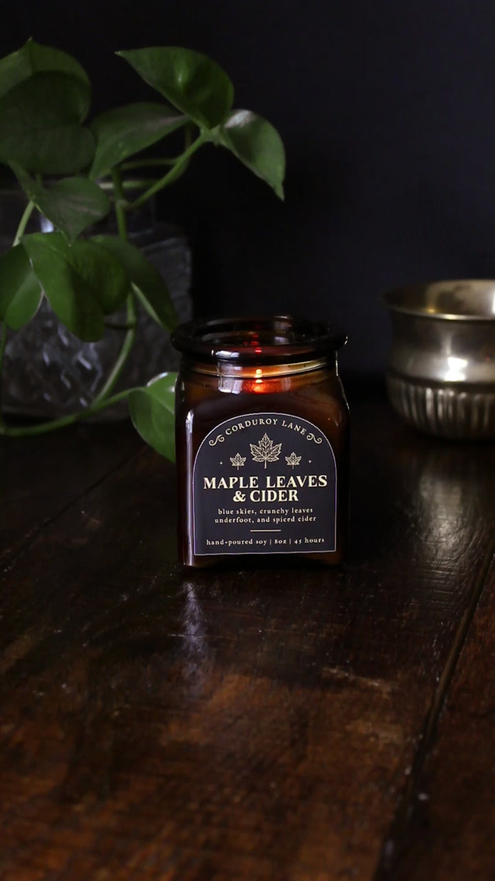 Maple Leaves and Cider: a cozy fall candle from Corduroy Lane in an amber glass square apothecary jar with a cork lid and a black and gold label, scented in a fresh, fruity blend of crunchy dried leaves and spiced apple cider for lovers of all things autumnal