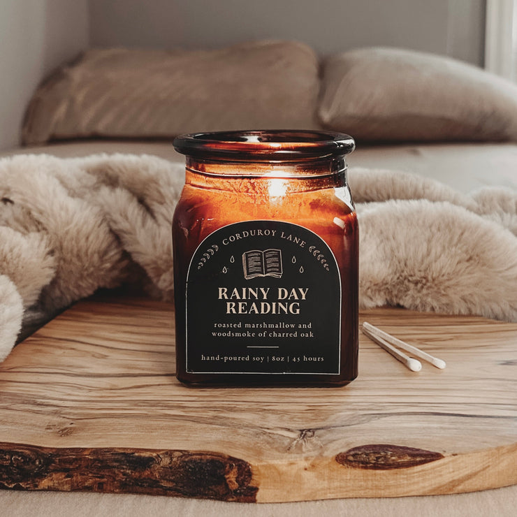 Rainy Day Reading: a cozy scented candle from Corduroy Lane. Get yourself in the mood to read with scents of toasted marshmallow, soft woodsmoke, and charred oak. An amber glass square jar with black and gold label and a cork lid in a black gift bag.